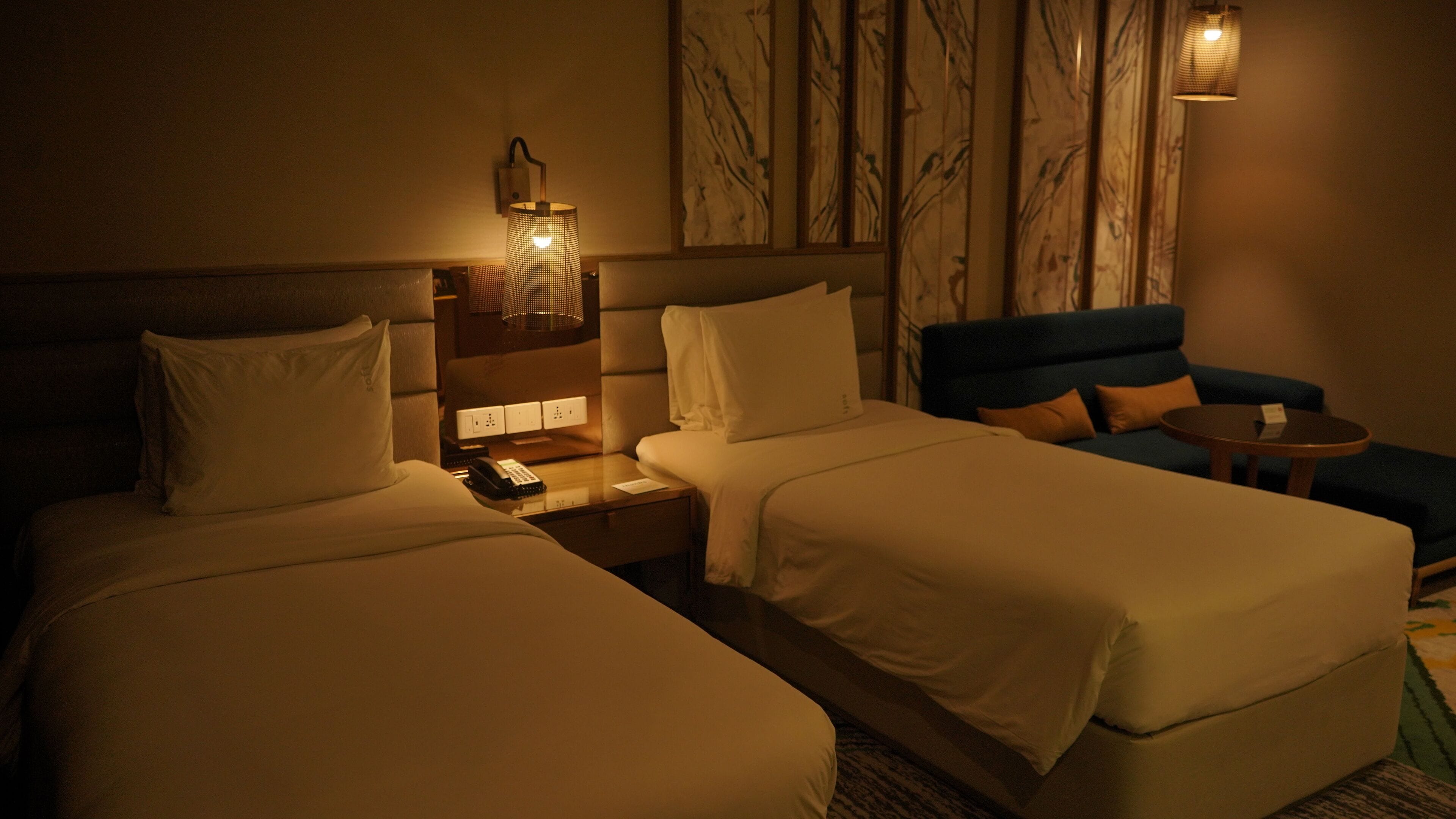 Una camera d'hotel con due letti, un comodino con lampada, un tavolino e un divano blu.
