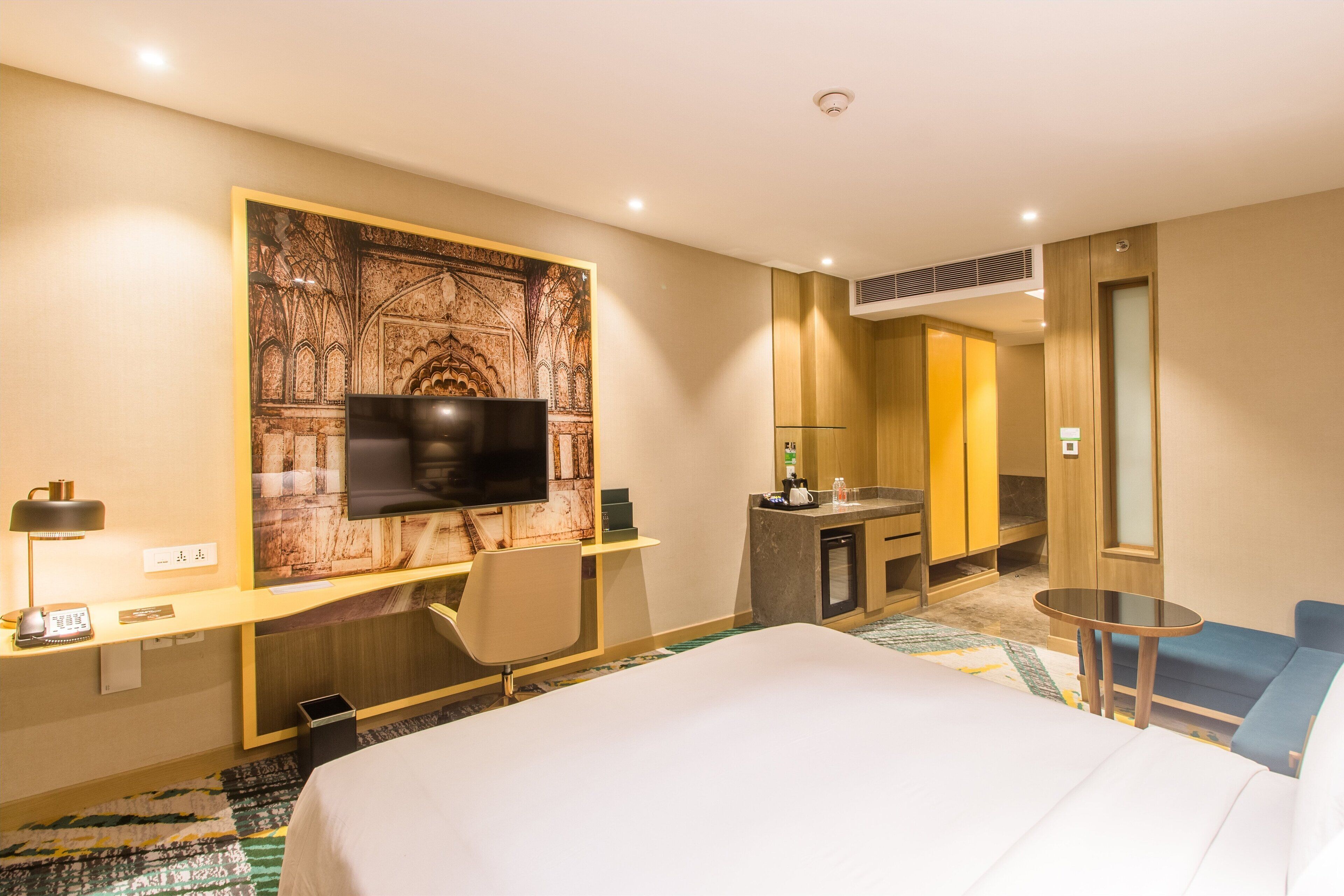 A hotel room with a large bed, a desk with a flat-screen TV, a small table, a chair, a bathroom with a mirror, and a wall-mounted artwork.