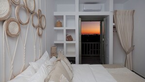 A bedroom with a bed, a white shelving unit, and a view of a sunset through an open door.