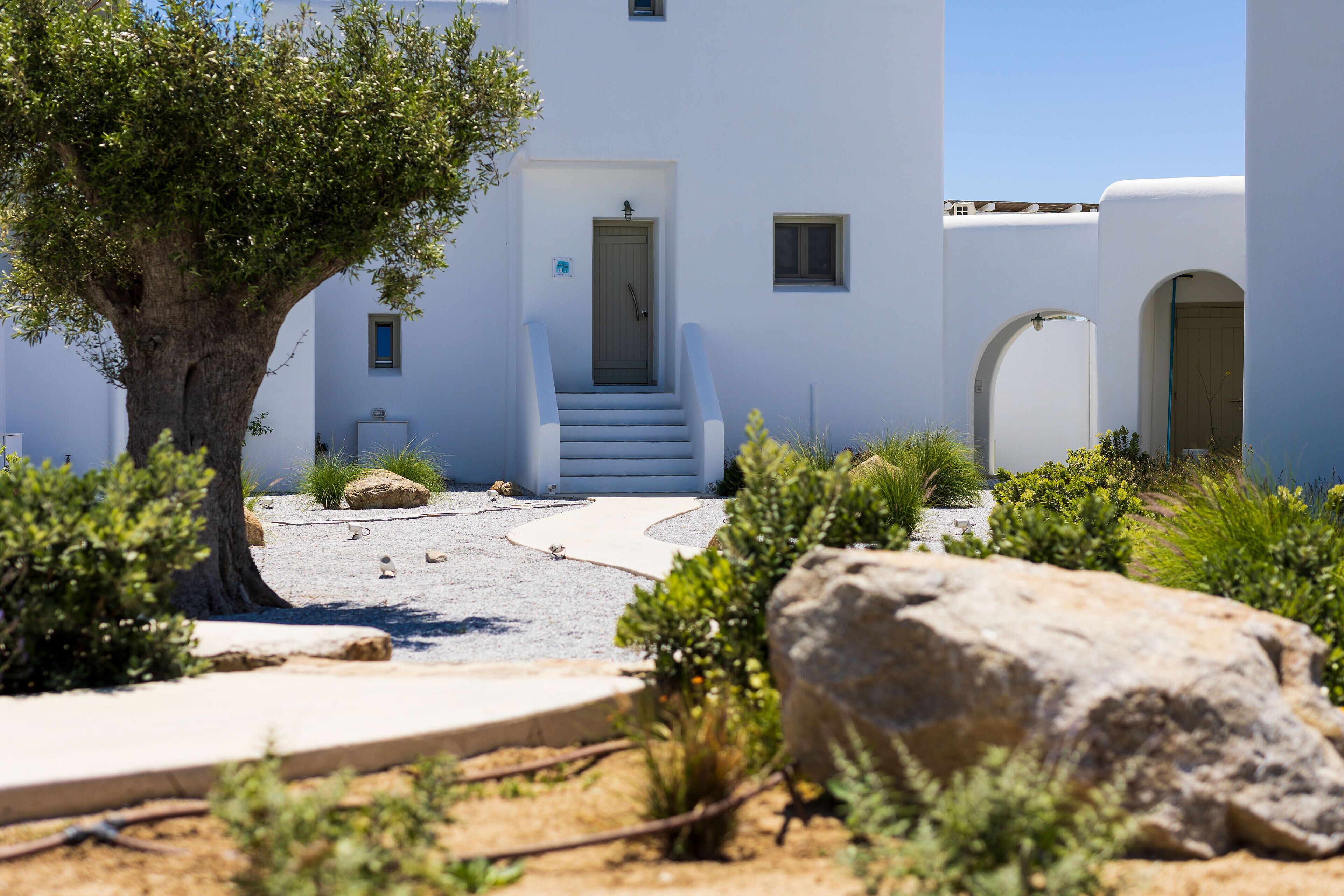 A white building with a tree, a large rock, and a staircase leading to a door.