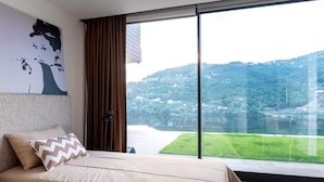 Quarto com uma janela grande, cama com roupa de cama bege, almofada com padrão em zigue-zague e um quadro na parede.