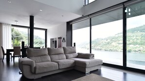 Uma sala de estar moderna com um amplo sofá em forma de L, uma área de refeições com mesa e cadeiras, e grandes janelas com vista para um lago e colinas.