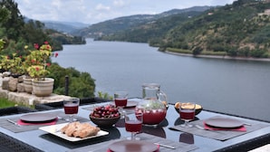 Disposição para refeições ao ar livre com vista panorâmica para um lago e colinas, incluindo uma mesa com pratos, copos, um jarro e uma tigela de cerejas.