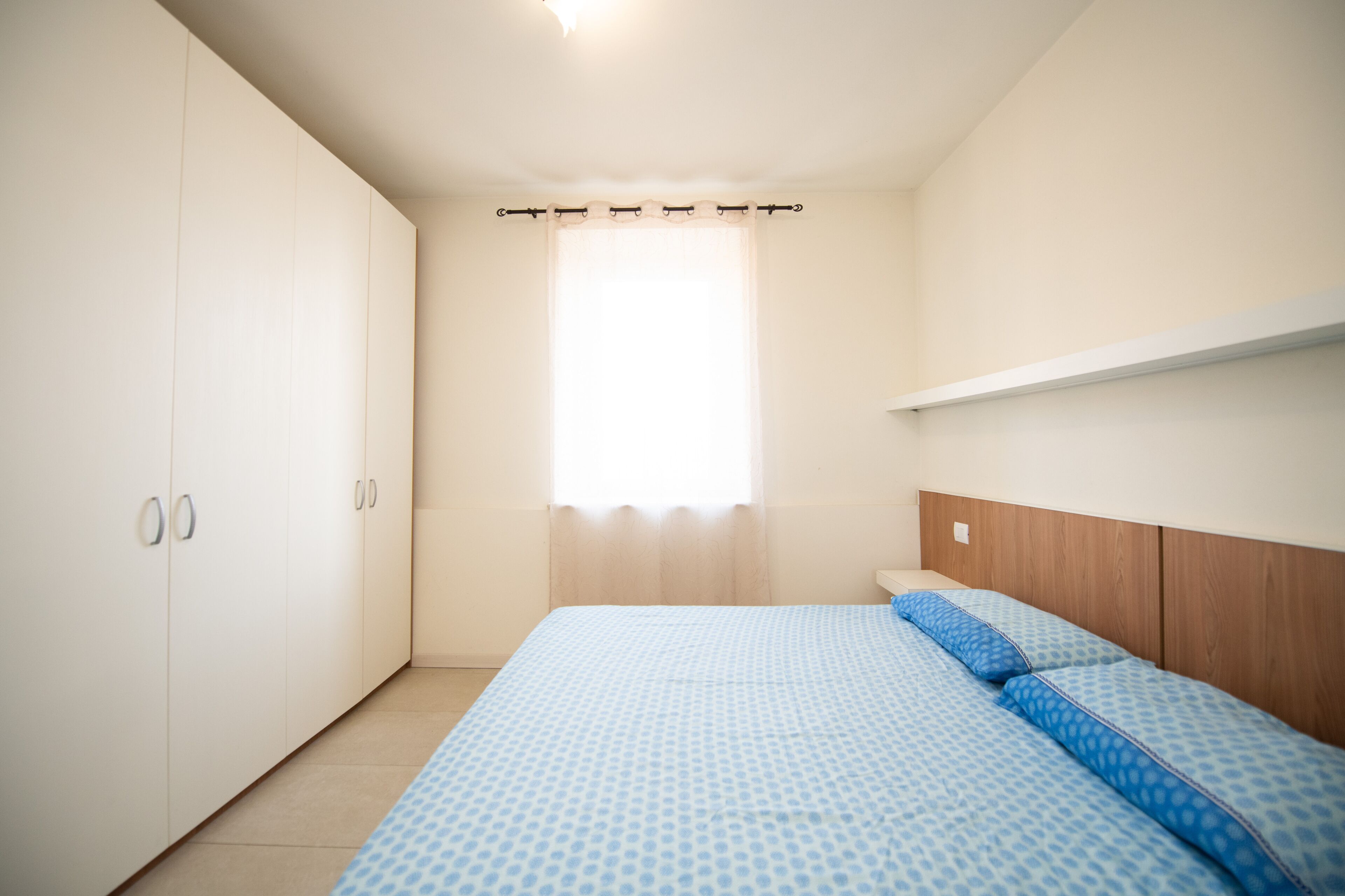 A bedroom with a bed, a wardrobe, a window with curtains, and a shelf above the bed.