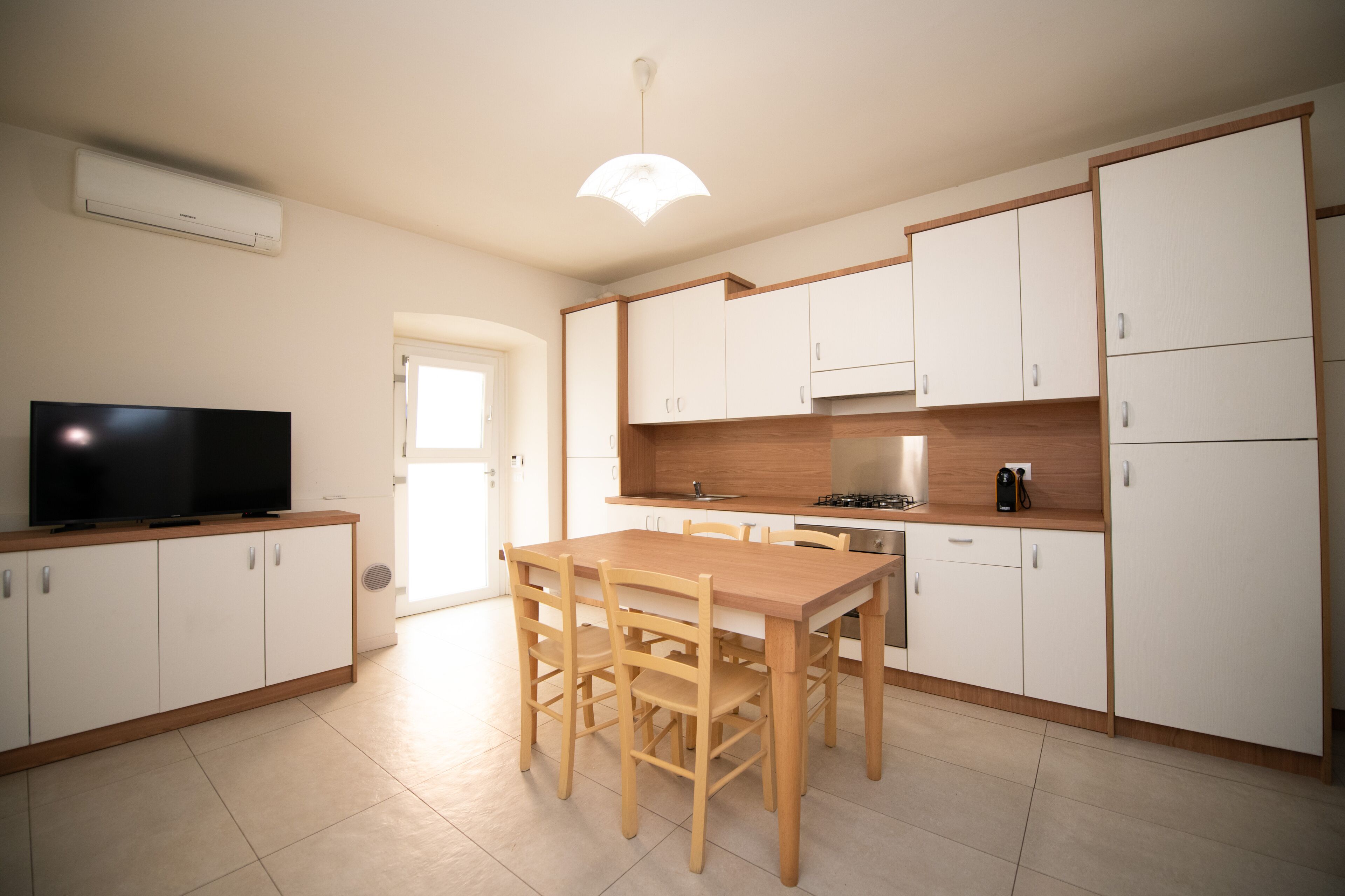 A modern kitchen with white cabinets, a wooden dining table, and a flat-screen TV.