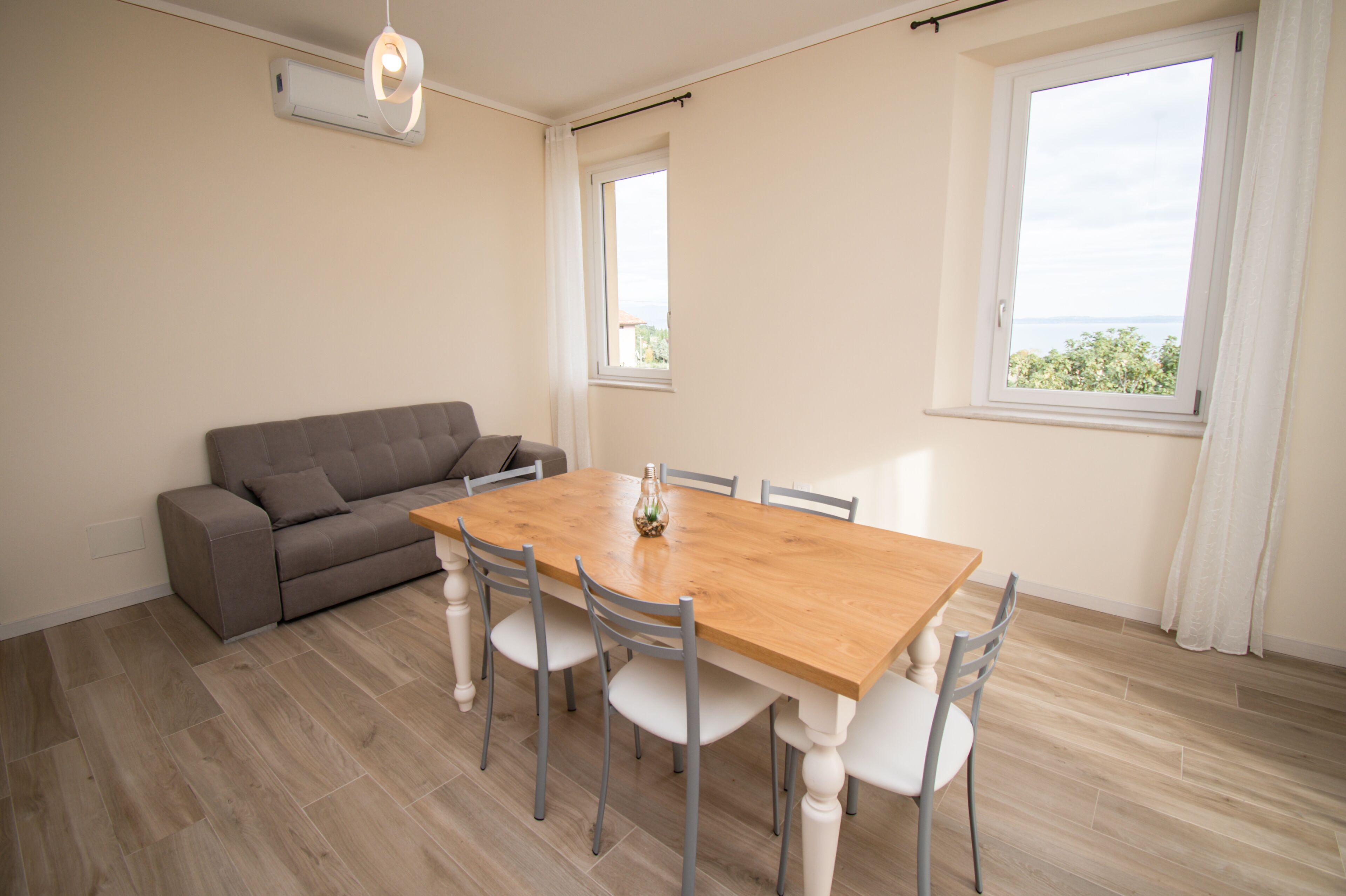 A dining area with a wooden table and chairs, a grey sofa, and a window with a view.