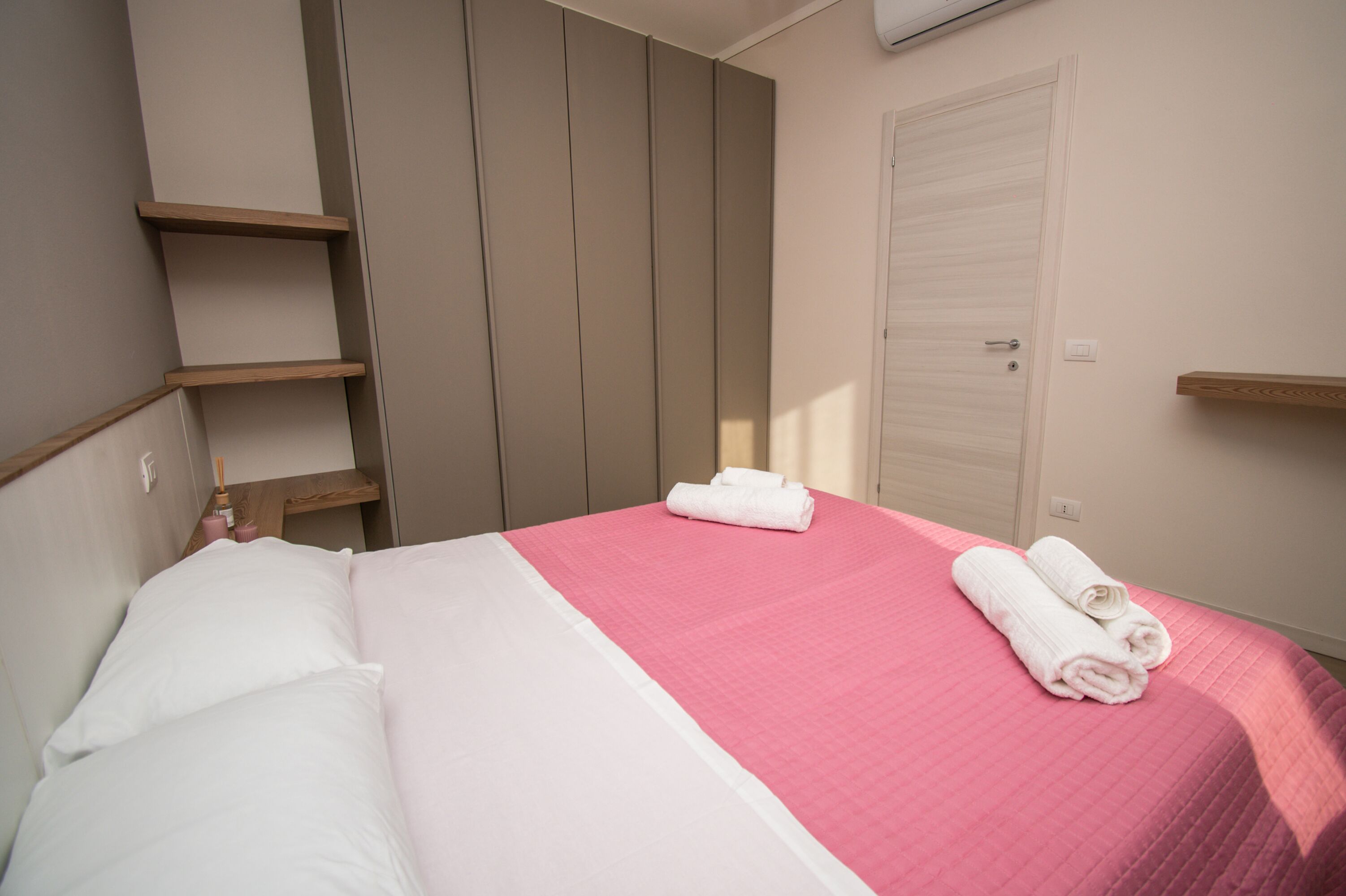 A neatly made bed with pink and white bedding, a wooden shelf, and a closed door.