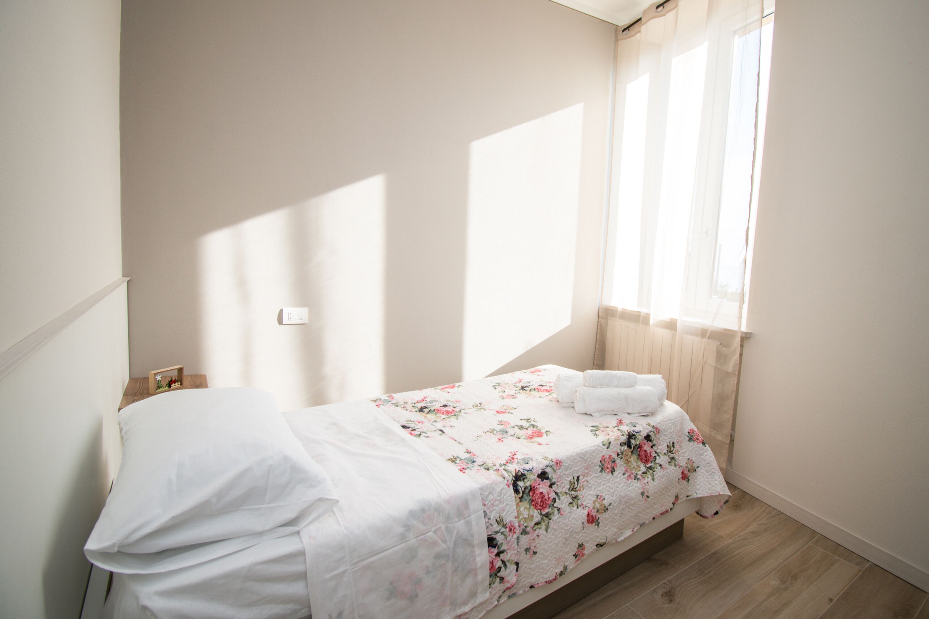A small, neatly made bed with a floral bedspread and white linens, a bedside table with a lamp, and a window with sheer curtains allowing natural light.