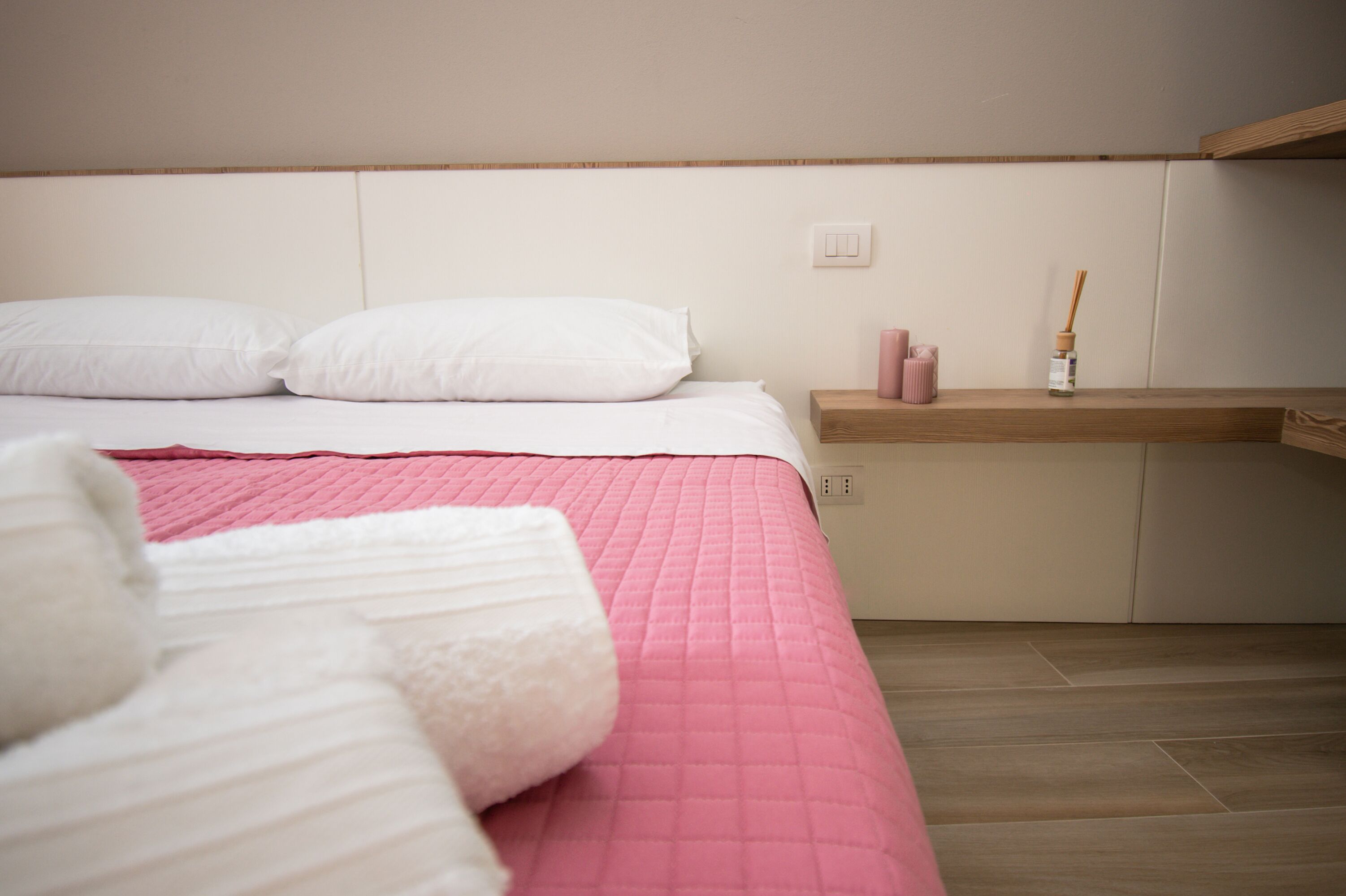 Una cama bien hecha con ropa de cama blanca y rosa, una mesita de noche de madera con una vela rosa y un difusor de caña, y un piso de madera.