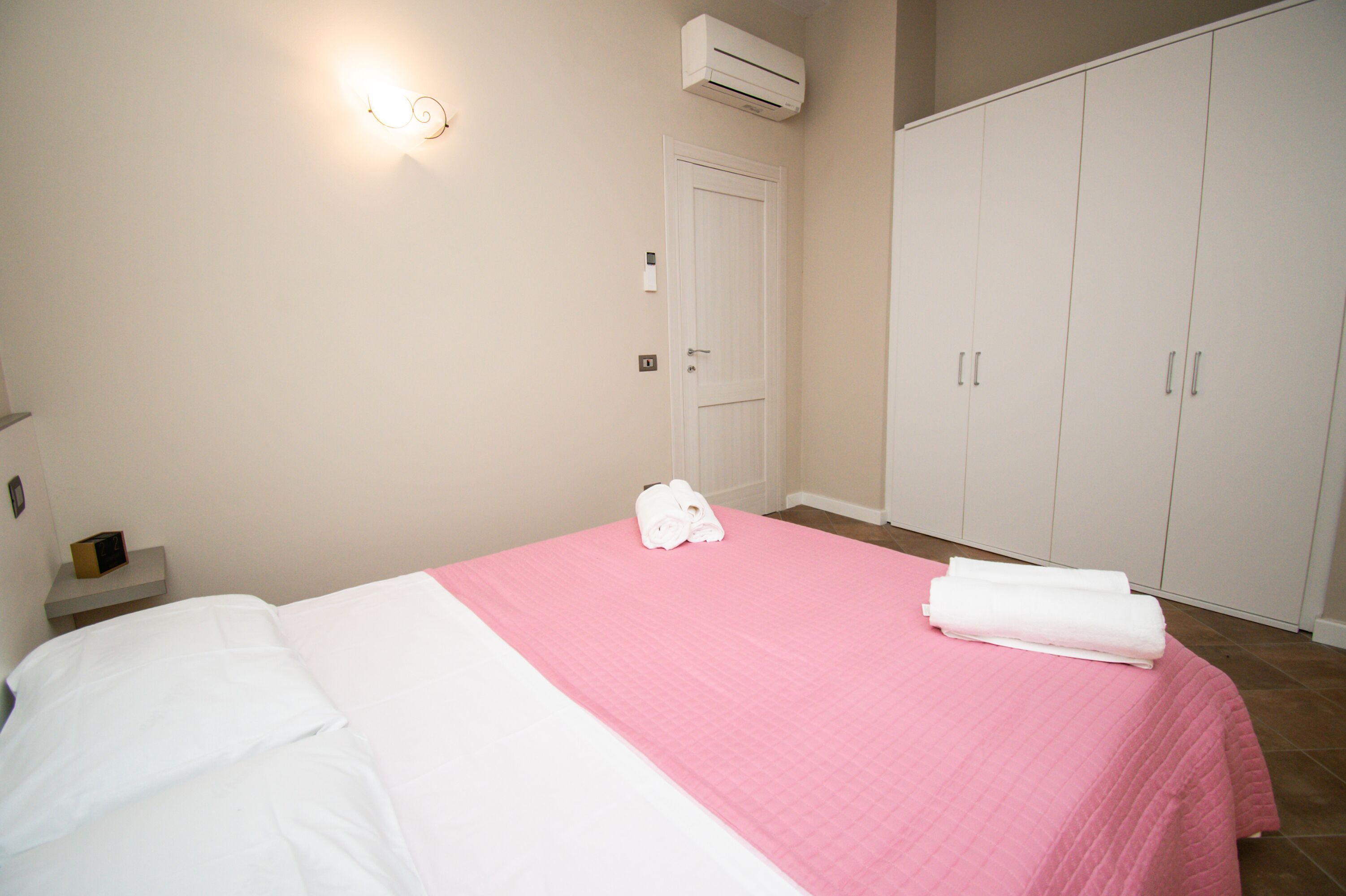 A bedroom with a bed, a pink bedspread, white pillows, a white towel, a nightstand, a wall-mounted light, a door, and a wardrobe.