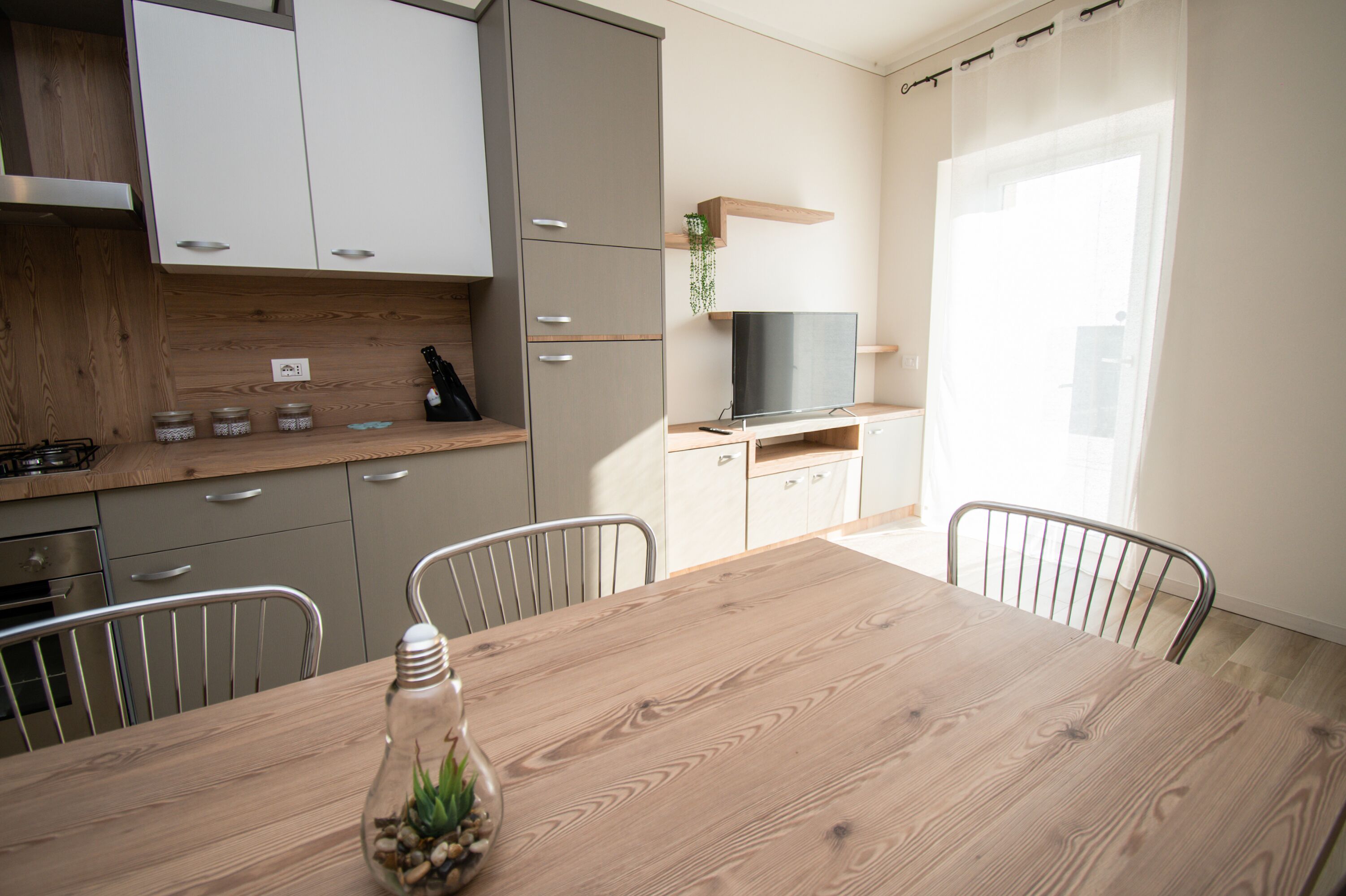 A modern kitchen with a wooden table, metal chairs, and a TV on a shelf.