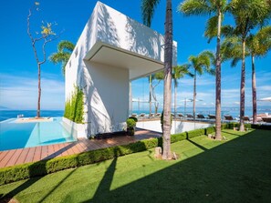 A modern building with a pool, surrounded by palm trees and a clear view of the sea.