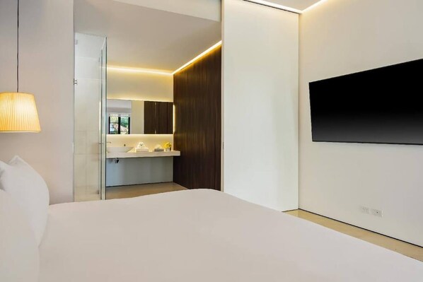 A modern hotel room with a bed, a TV mounted on the wall, a bedside lamp, and a bathroom visible through a glass door.