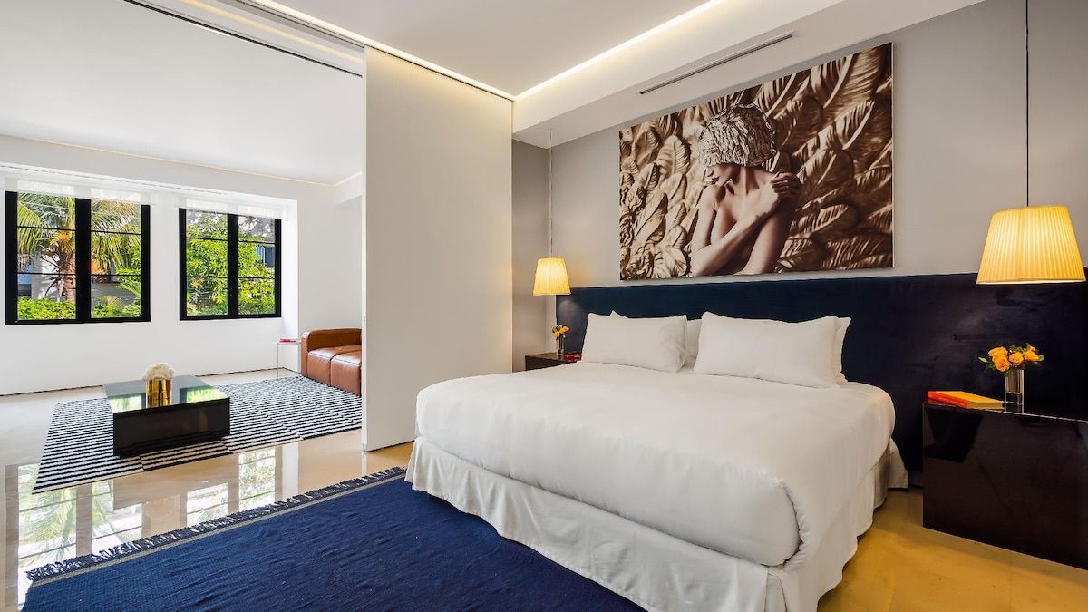 A modern hotel room with a large bed, two bedside lamps, a wall-mounted artwork, and a view through large windows.