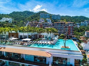 A rooftop swimming pool with sun loungers, surrounded by palm trees and modern buildings.