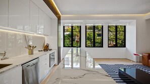 A modern kitchen with a marble countertop, built-in appliances, and large windows offering a view of greenery.