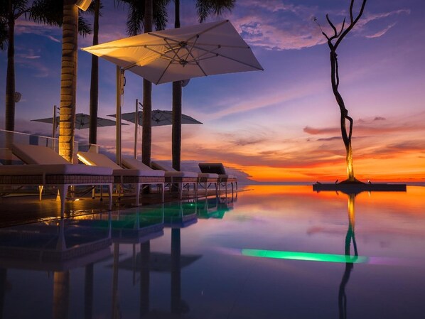 An infinity pool with lounge chairs and umbrellas, reflecting a sunset over water.