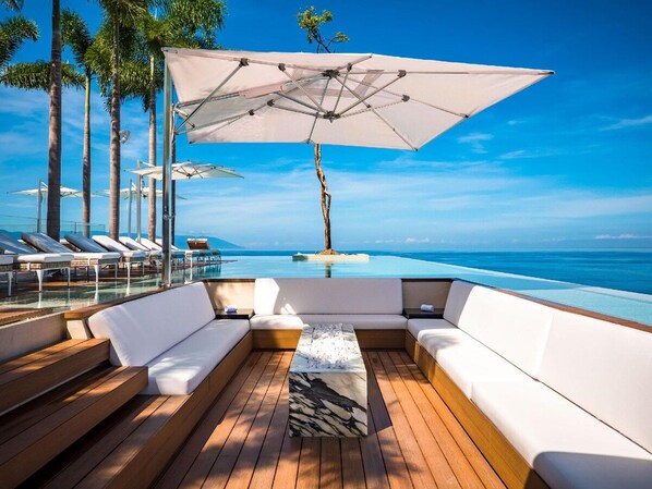Zona de piscina con terraza de madera, asientos blancos acolchados y un paraguas grande.