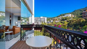 Un balcón con una mesa y sillas blancas, con vistas a una colina con edificios y vegetación.