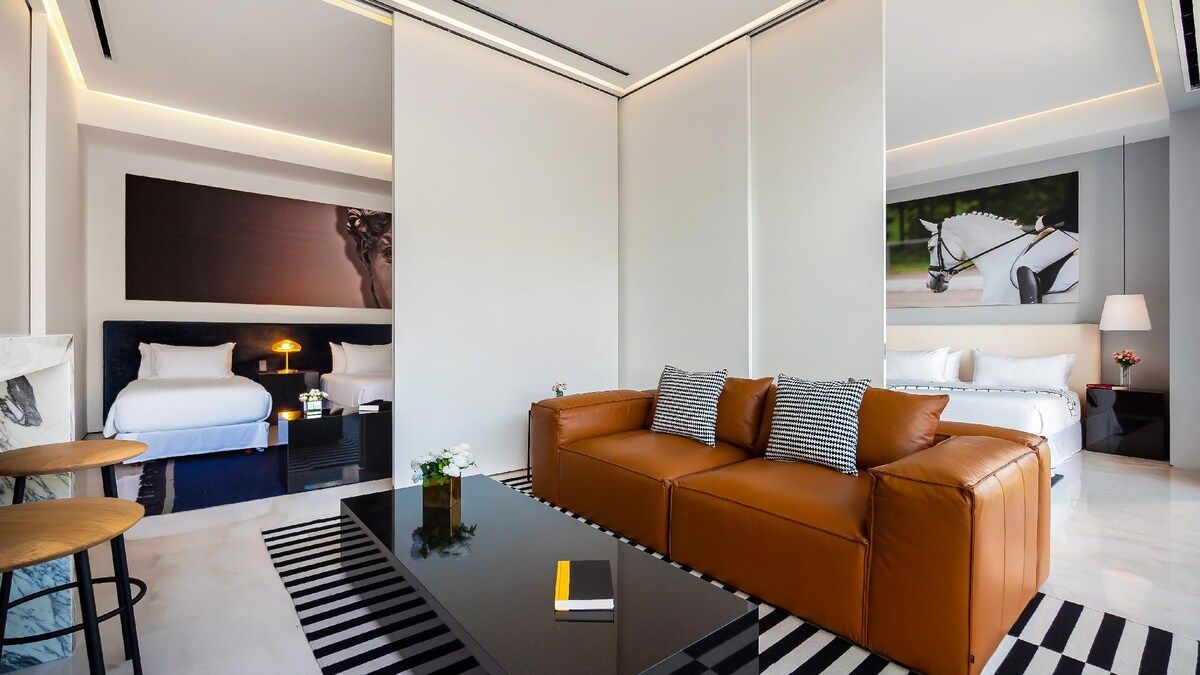 Une chambre d’hôtel moderne avec un canapé en cuir, un lit avec du linge de maison blanc, une table de chevet avec une lampe et un tableau au mur.