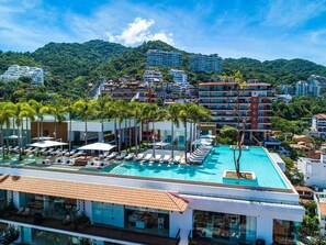 A rooftop swimming pool with sun loungers and palm trees, surrounded by buildings and a mountainous backdrop.