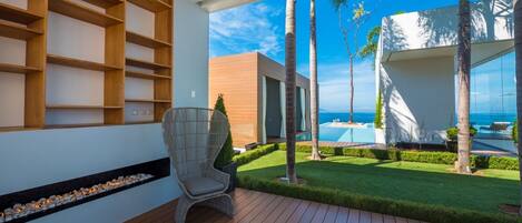 Un espacio exterior moderno con terraza de madera, una silla de mimbre y vistas a una piscina y palmeras.