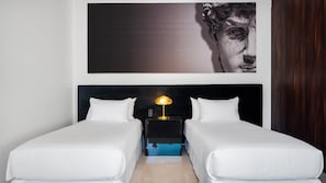 Habitación de hotel con dos camas individuales, una mesita de noche con lámpara y un cuadro abstracto grande sobre las camas.