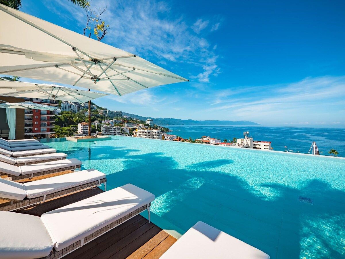 En infinitypool med vita solstolar och ett vitt parasoll, med utsikt över en kuststad.