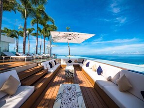A sunlit deck with wooden flooring, white sofas with cushions, a central coffee table, and a large umbrella providing shade.