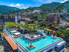 A rooftop pool with lounge chairs and palm trees, surrounded by buildings and a mountainous backdrop.