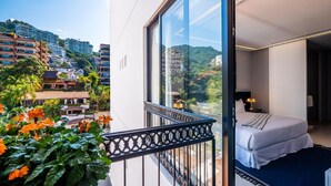 Un balcón con vistas a un paisaje urbano, una cama con colchón blanco y un detalle en azul, y una mesita con una lámpara.