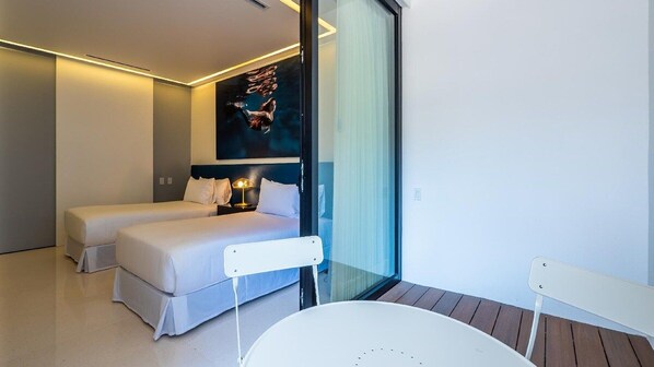 A modern hotel room with two beds, a glass partition, a small table, and a view of the outdoors.