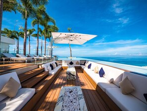 A sunlit deck with wooden flooring, white sofas, and a central table, overlooking a clear blue ocean.