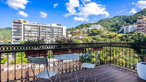 Un balcón con vistas a una colina, un edificio al fondo y una mesa con sillas.