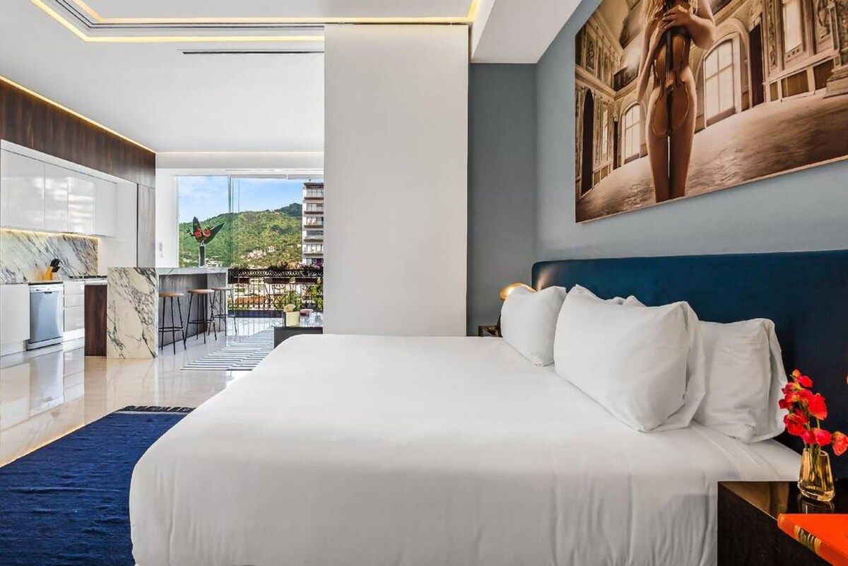 A modern hotel room with a large bed, a view of a cityscape, and a dining area.