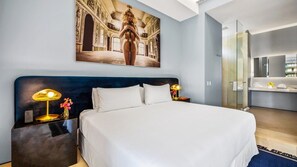 Habitación de hotel con una cama grande, dos lámparas de noche, un espejo y un baño con cabina de ducha de vidrio.