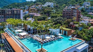Una piscina en la azotea con sillones de descanso y palmeras, rodeada de edificios y un paisaje montañoso.