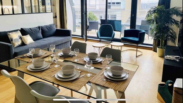 A modern living room with a glass dining table set for four, a sofa, and a balcony with chairs. - Luxury Green Penthouse Center - Parking (Luxembourg City)