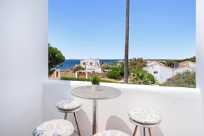 A balcony with a table and two stools, overlooking a coastal town with buildings and a clear sky. - Charming Duplex with Sea Views, 2 Bedrooms + 3 Bathrooms, 3 Minutes to Beach (Arenal d'en Castell, Es Mercadal)