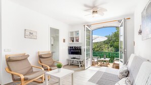 Un salón luminoso con un sofá, dos sillones, una mesita de centro y un televisor. Hay un balcón con vistas a zonas verdes.