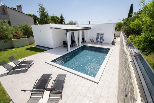 Ein rechteckiger Swimmingpool mit weißer Liegefläche, umgeben von einem Holzzaun. Liegestühle und eine überdachte Sitzgruppe mit Tisch und Stühlen sind vorhanden.