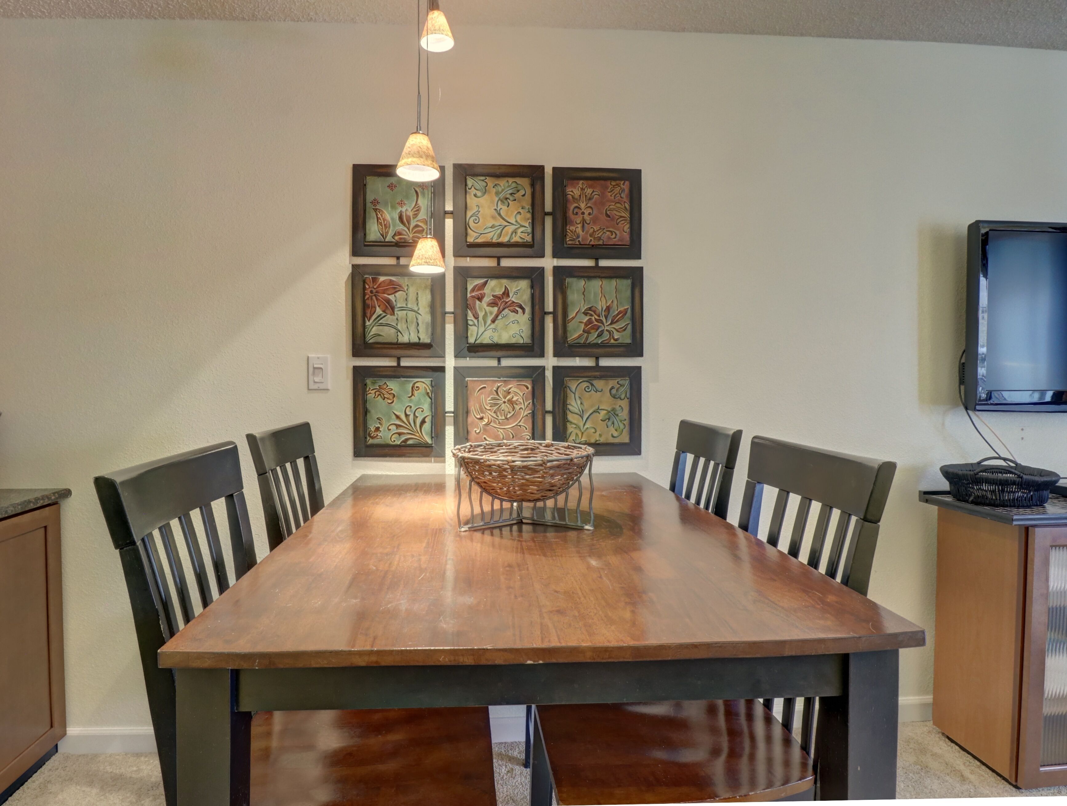 A dining area with a wooden table, six chairs, a woven basket, and framed floral artwork on the wall.