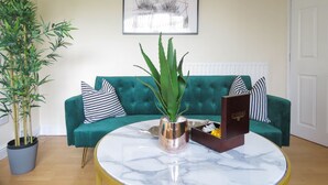 Ein Wohnzimmer mit einem salbeigreenen Sofa, einem runden Marmortisch, einer Topfpflanze und einem gerahmten Bild an der Wand.
