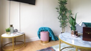 Ein Wohnzimmer mit Flachbildfernseher an der Wand, einem kleinen runden Tisch mit einer Zeitschrift, einem pinken Pouf, einer grünen Pflanze und einer blauen Decke.