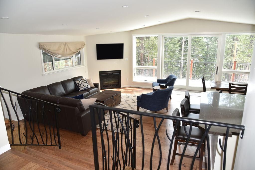 Una sala de estar con chimenea, un sofá, una zona de comedor con mesa y sillas, y ventanas grandes con vistas a árboles.
