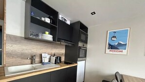 A compact kitchen with a wooden countertop, black cabinets, a built-in microwave, and a small sink. A chair is placed near the sink, and a framed picture of a mountain and cable car is on the wall. - Apt, 1206 _ Properties Mont Amour  (Morzine)