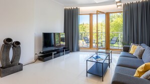 Een moderne woonkamer met een flatscreen televisie, een bank, een salontafel en een tafel met glazen blad. Er is een groot raam met gordijnen en er is een balkon zichtbaar door het raam.
