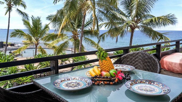 Una mesa con un arreglo de frutas tropicales, dos platos estampados y una silla de mimbre con cojín.