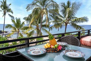 Una mesa con un arreglo de frutas tropicales, dos platos estampados y una silla de mimbre con cojín.