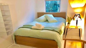 Un lit bien fait, avec une couverture verte et des oreillers, une tête de lit en bois, une petite table de chevet avec une lampe, une chaise et une fenêtre donnant sur des arbres.